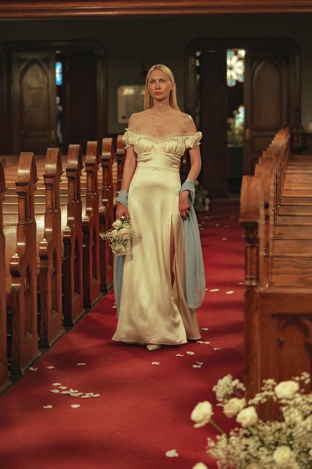 Model walks down the aisle in an off-the-shoulder ivory bridal gown during Tanner Fletcher's bridal fashion show at New York Bridal Fashion Week, carrying a bouquet inside a historic church adorned with rose petals.